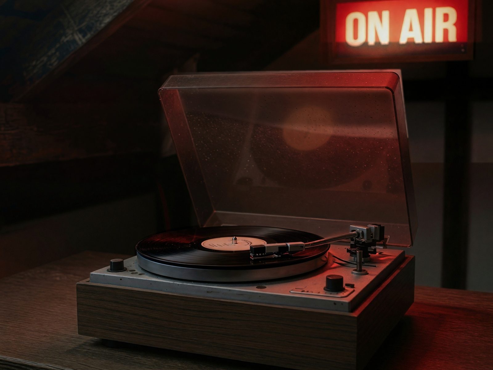 A turntable with its smoked lid up, on a wooden table under a lit ON AIR sign.